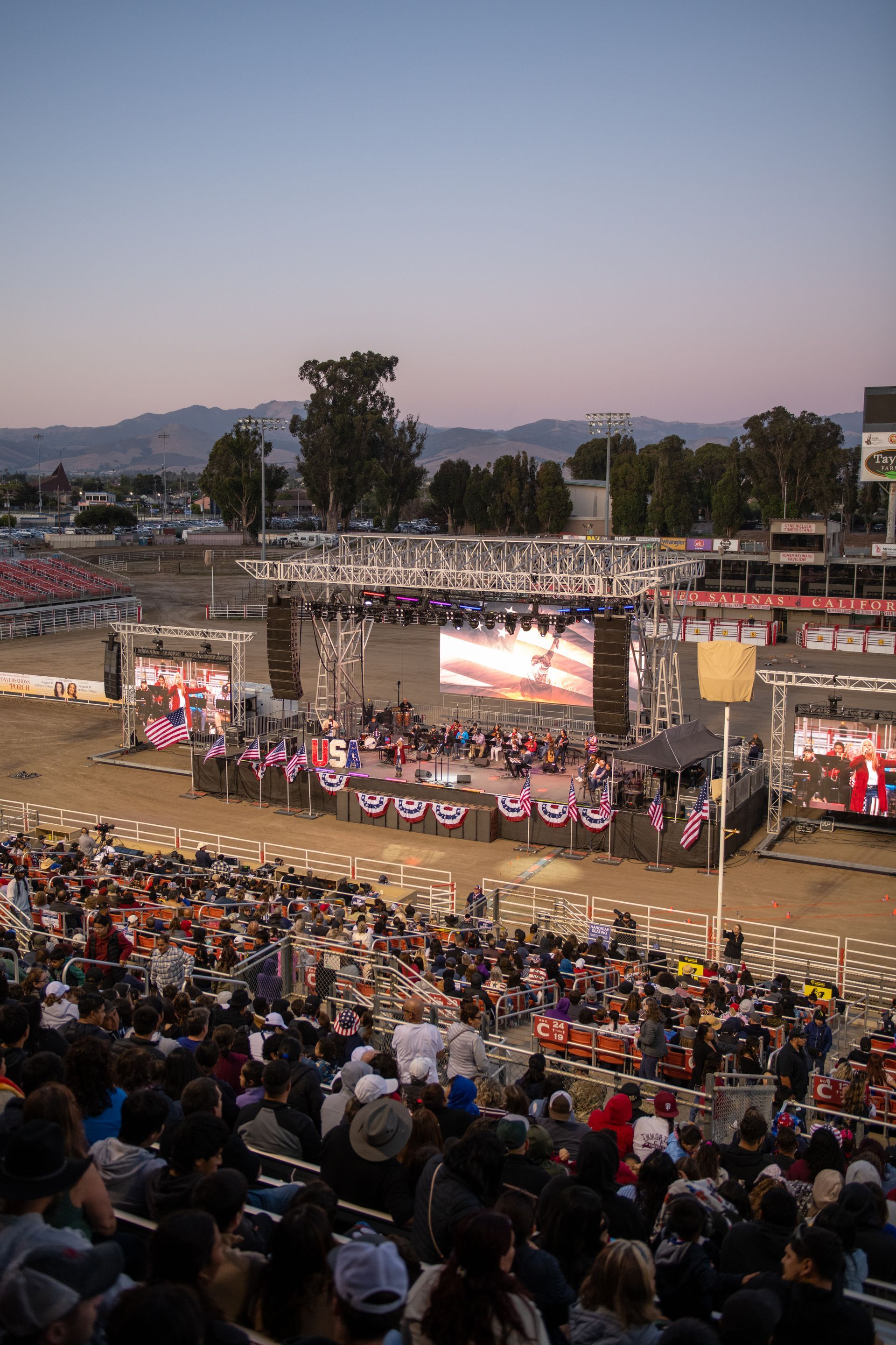 Large outdoor event with stage and screens; crowd in bleachers.