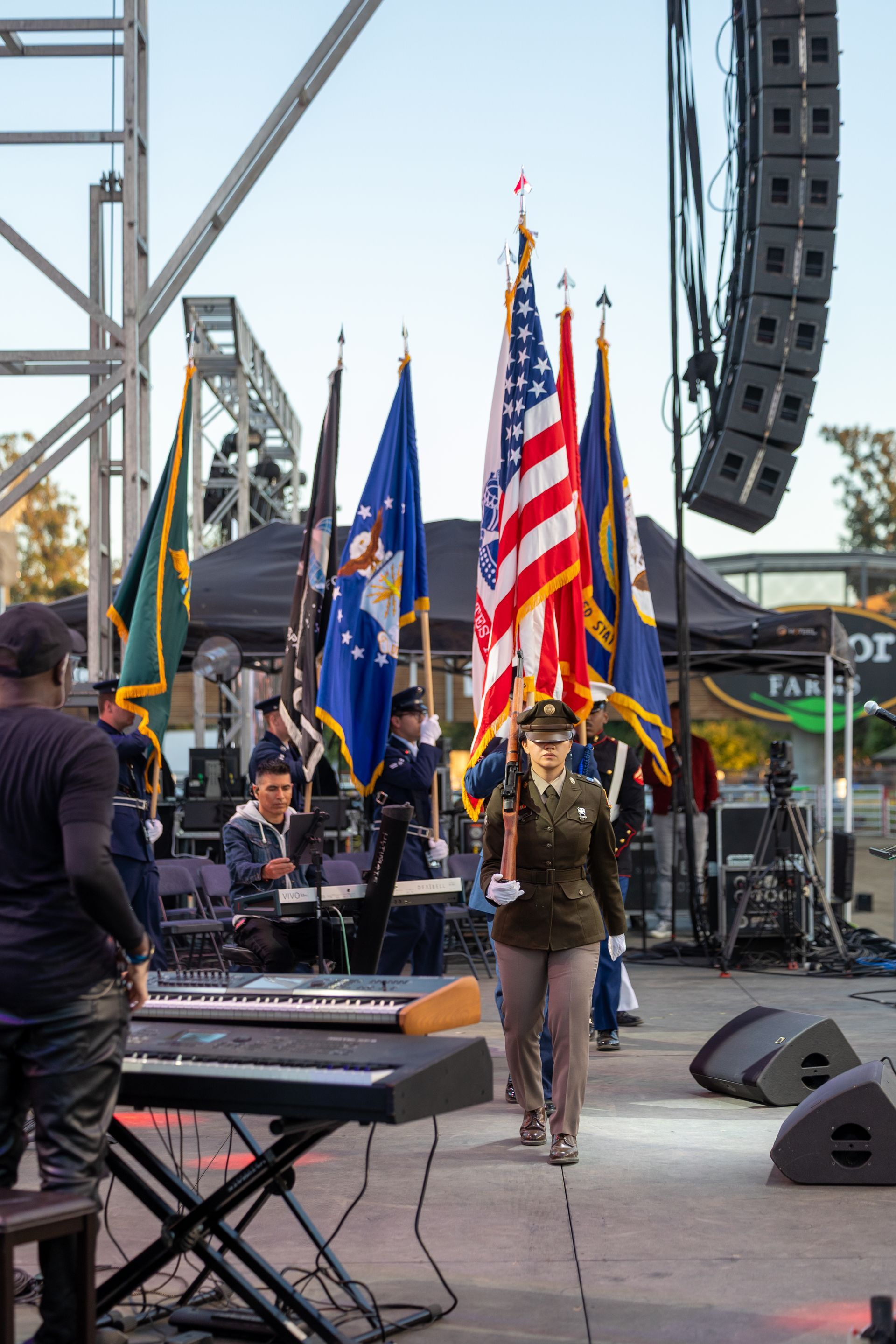 Soldier in uniform marches with flags; band plays on outdoor stage.