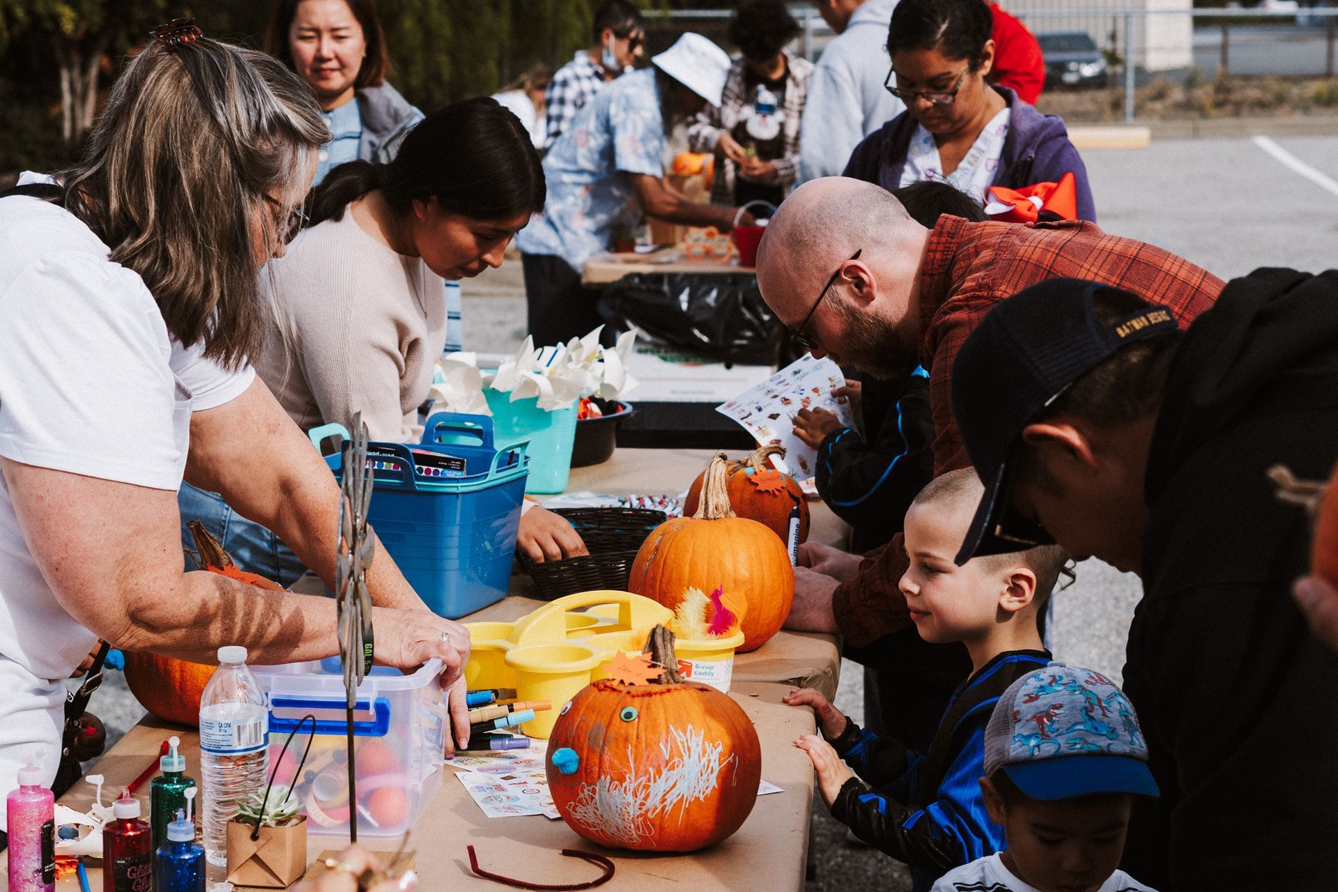 People decorate pumpkins at an outdoor event, using craft supplies at a table.