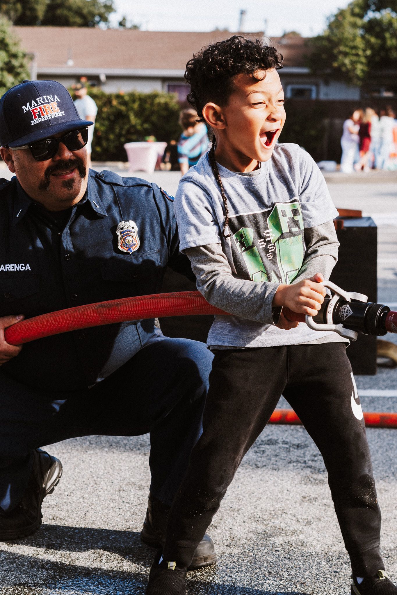 A firefighter helps a child hold a hose, both smiling outdoors.