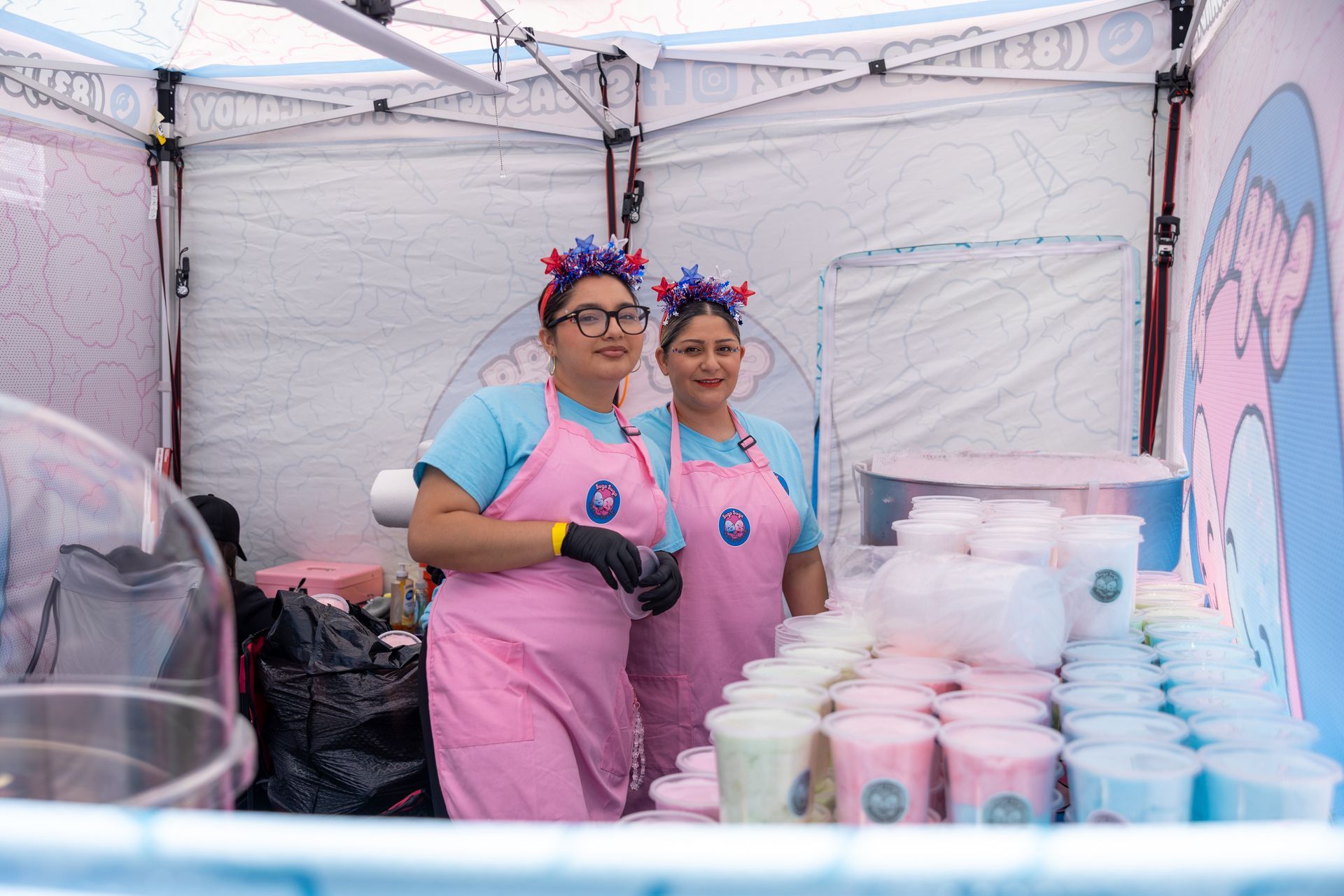 Two people in pink shirts and aprons stand behind a table of cotton candy at an outdoor booth.