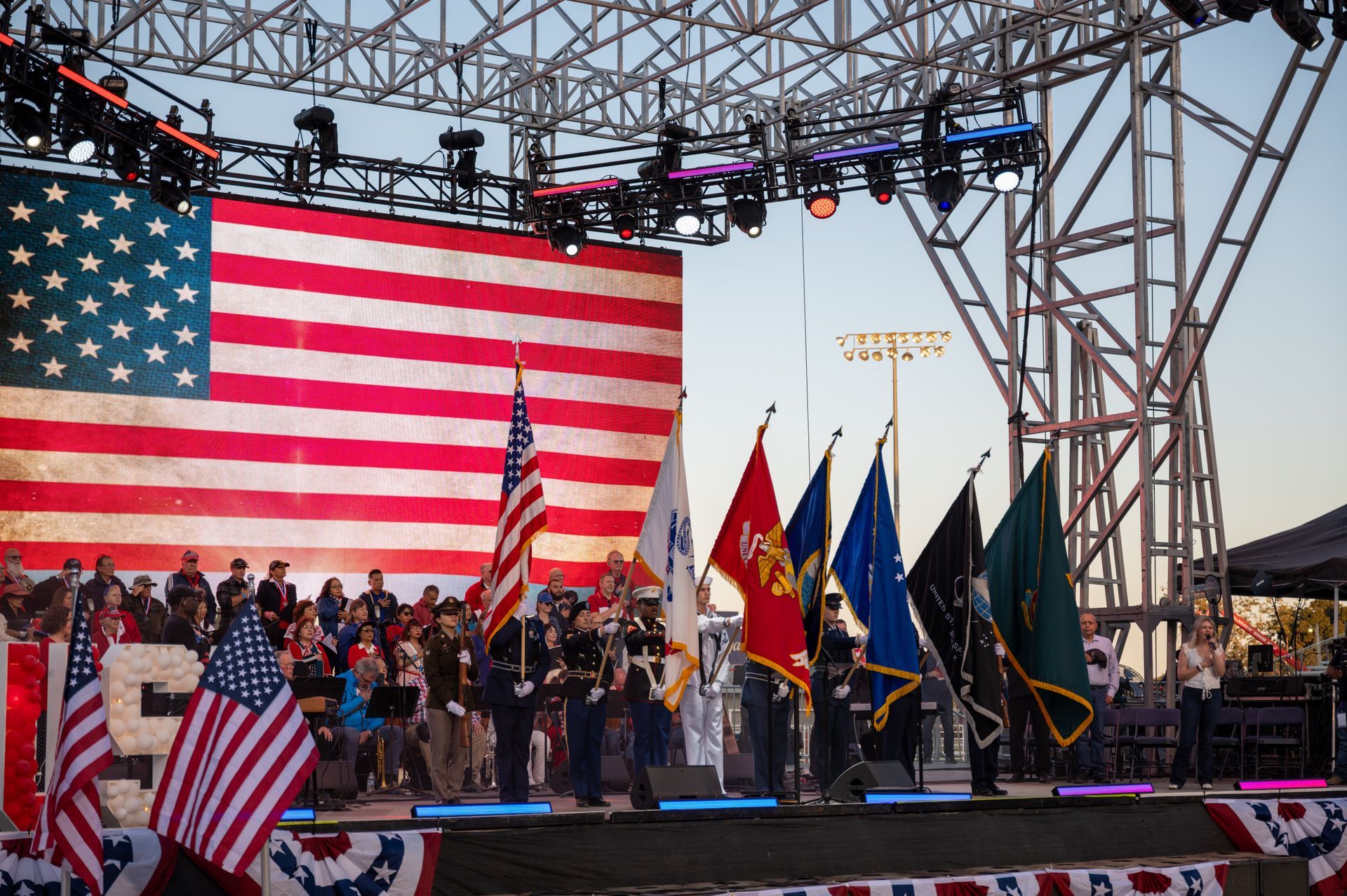 Crowd on stage with U.S. flag backdrop and military-style flags at a patriotic outdoor event