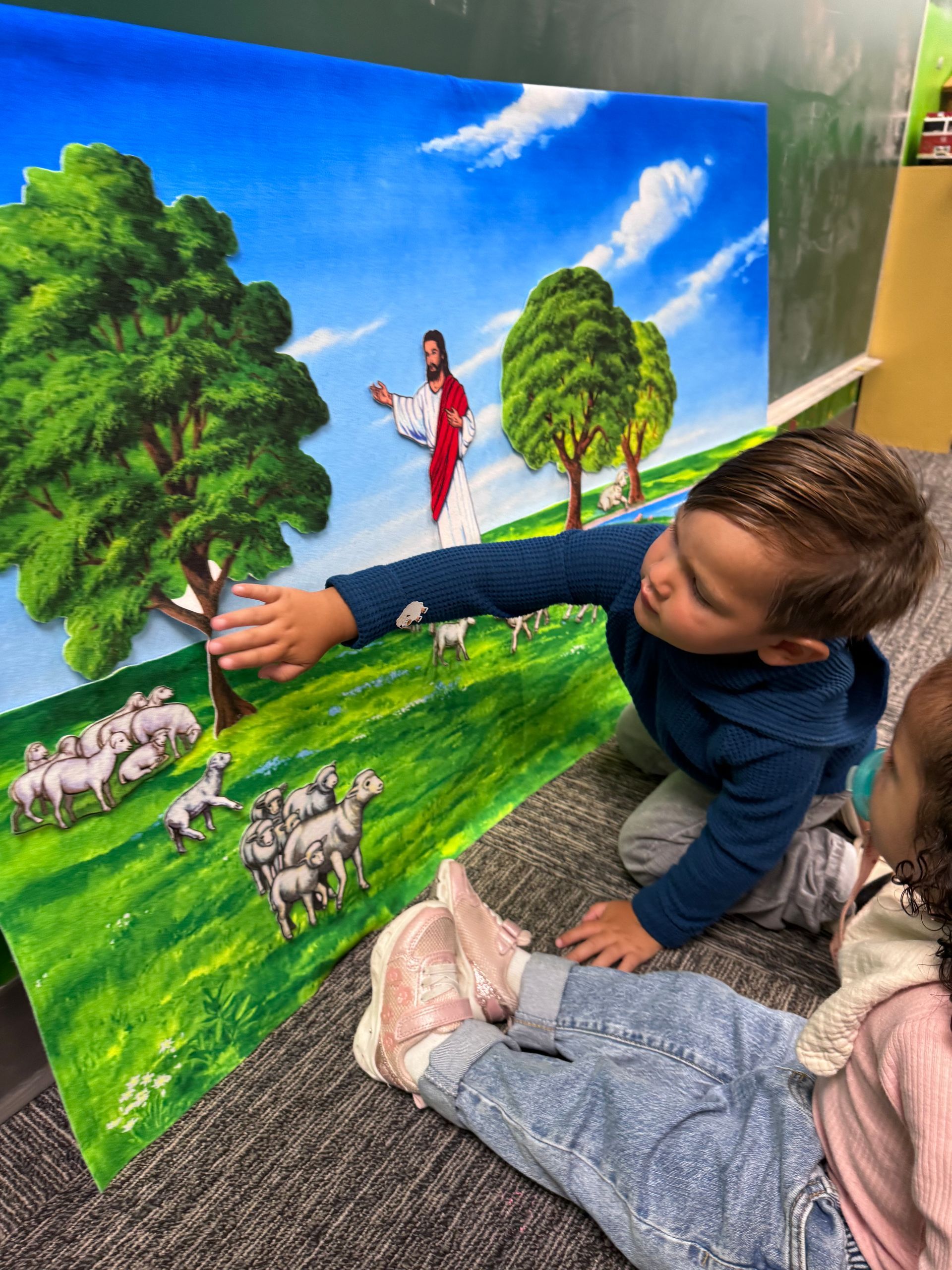 A child is pointing at a colorful painting of trees, sheep, and a figure in a red robe outdoors