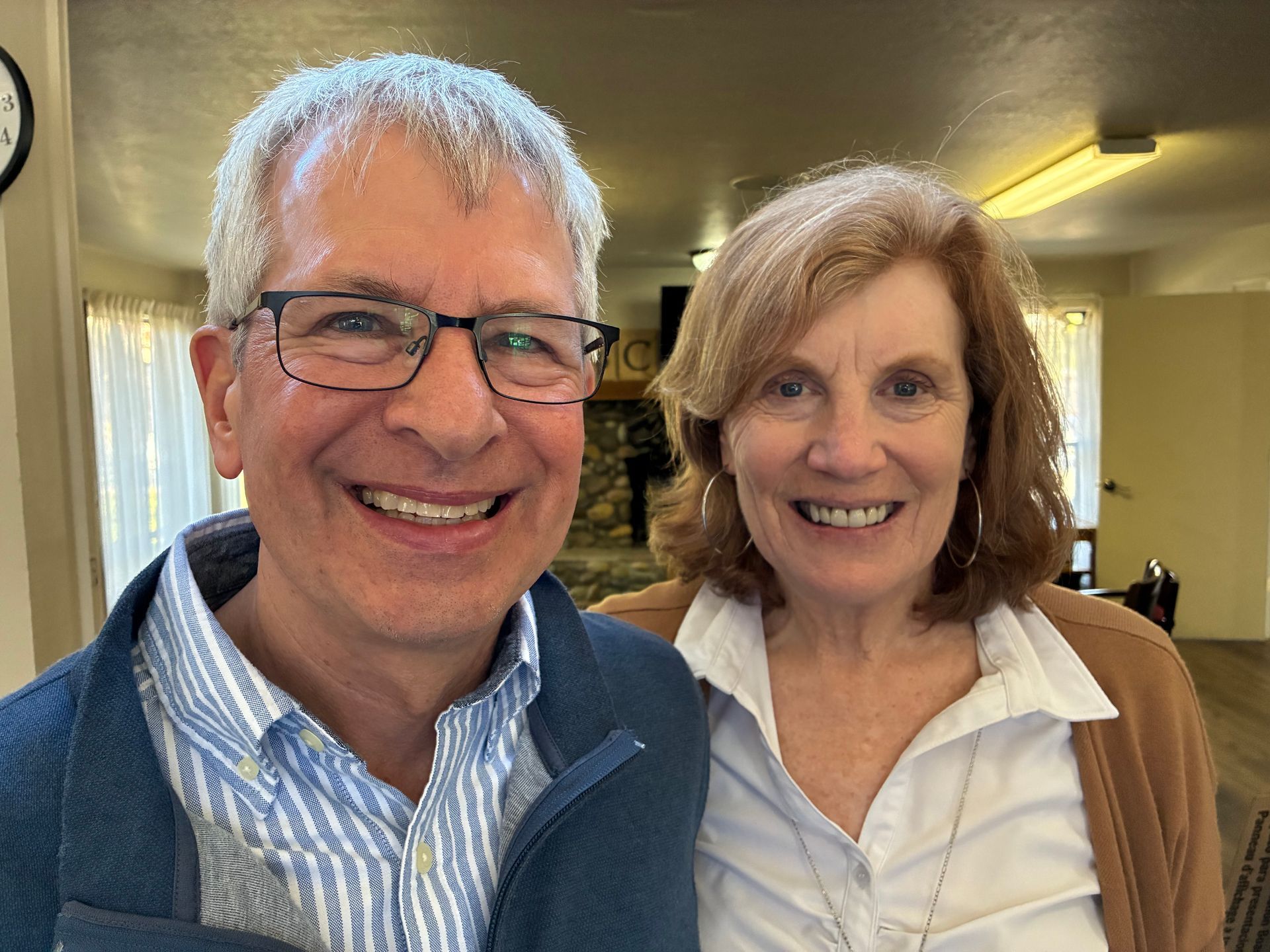 Two smiling people indoors, wearing glasses and casual sweaters in a hallway.