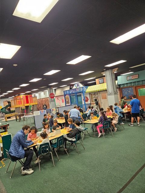 Children sit at tables in a colorful indoor play space with carpet and bright ceiling lights.