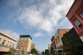 Buildings line a city street, viewed angled upwards toward a blue sky with clouds.