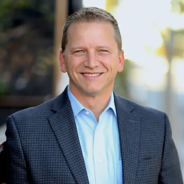 Man in a suit smiles, outdoors. He wears a blue button-down shirt under a gray blazer.