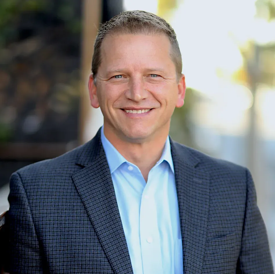 Man in blue shirt and blazer smiles outdoors.