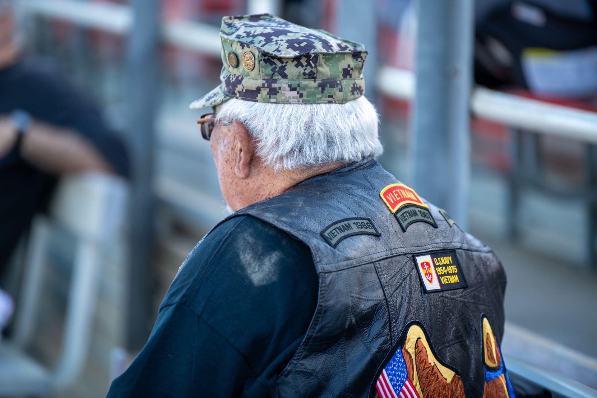 Veteran in camouflage cap and vest, seen from behind in stadium seating with a small U.S. flag patch
