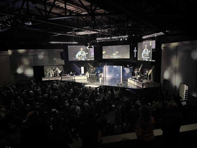 Large auditorium with a stage; people watching a performance with screens, lights, and musicians.