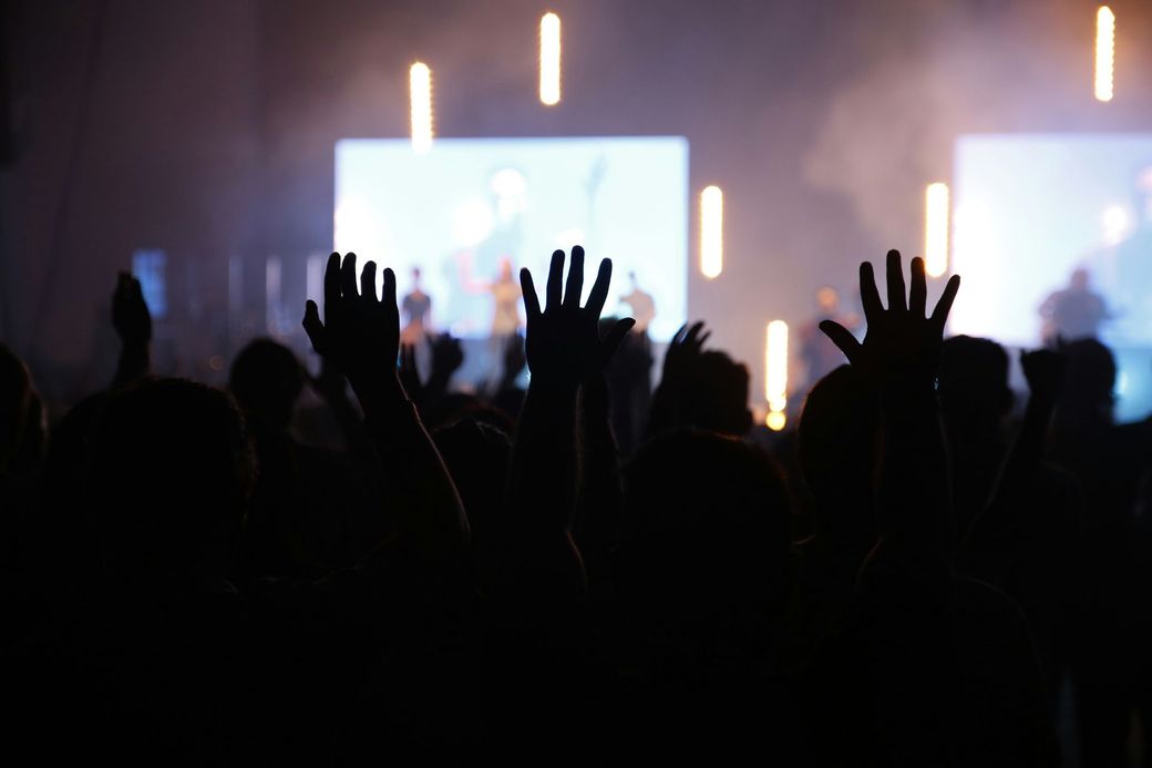 Audience raises hands in worship during a performance with stage lights and screens.
