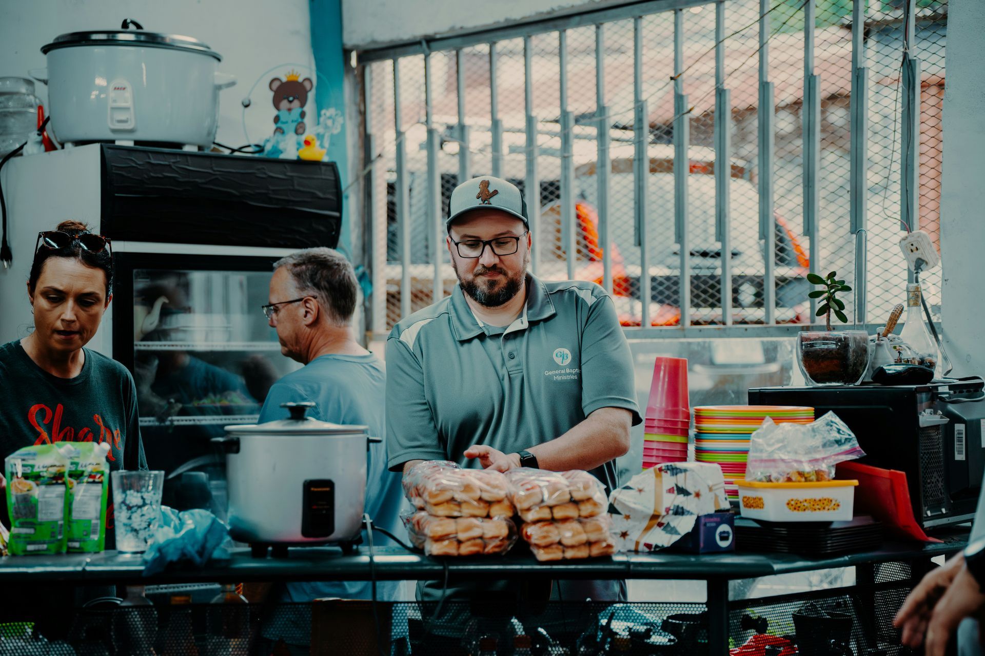 People behind a counter at a food stall. One man in a hat and grey shirt, looking at camera. Stacks of bread and cooked food.