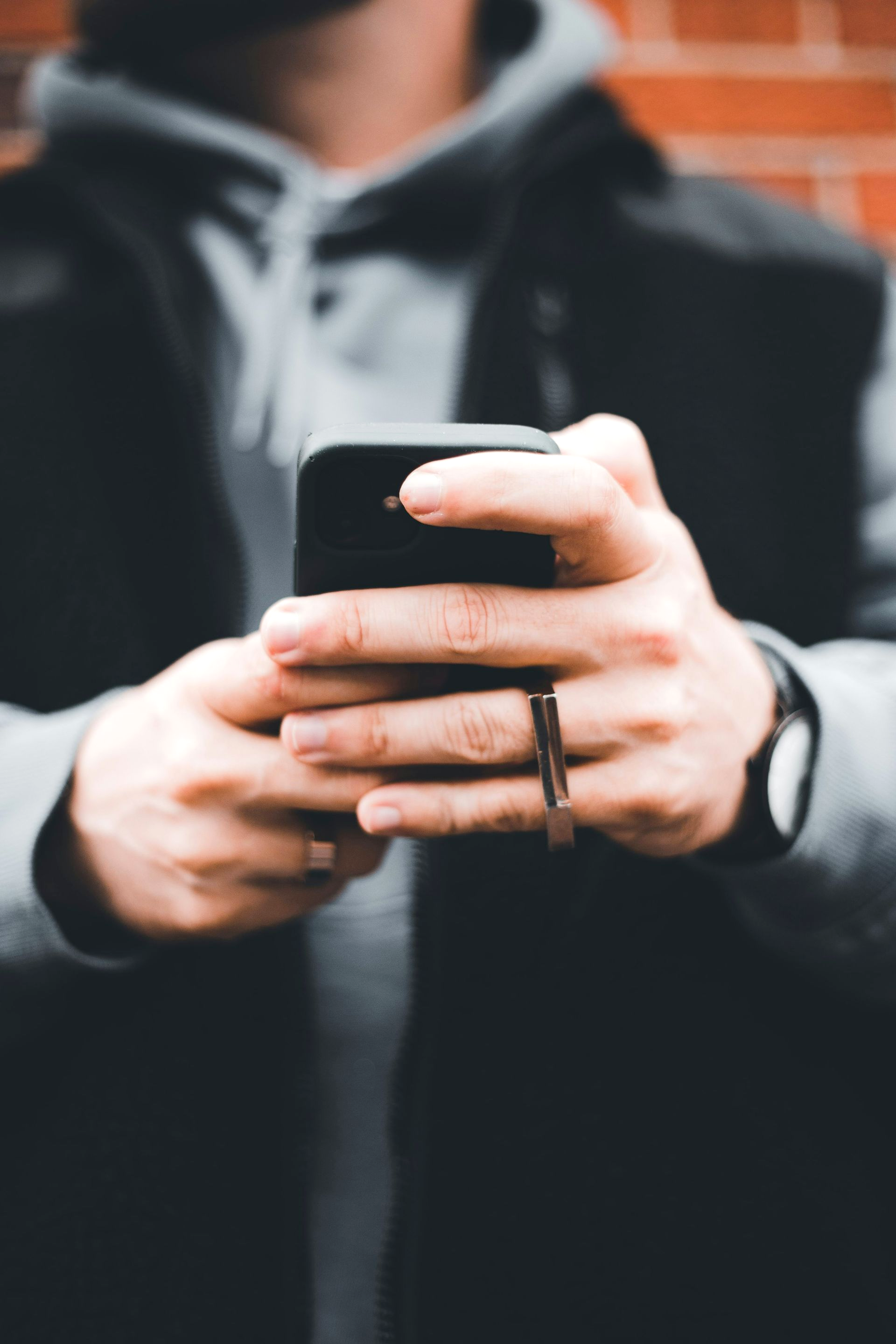 Man holding a black smartphone, wearing a ring, jacket, and hoodie, against a brick wall.