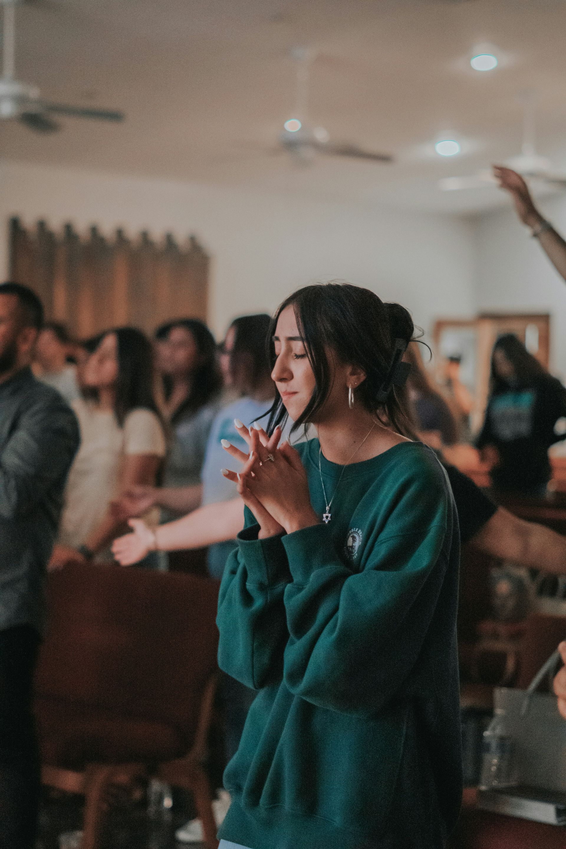 Woman with hands clasped, eyes closed, praying in a group of people indoors.