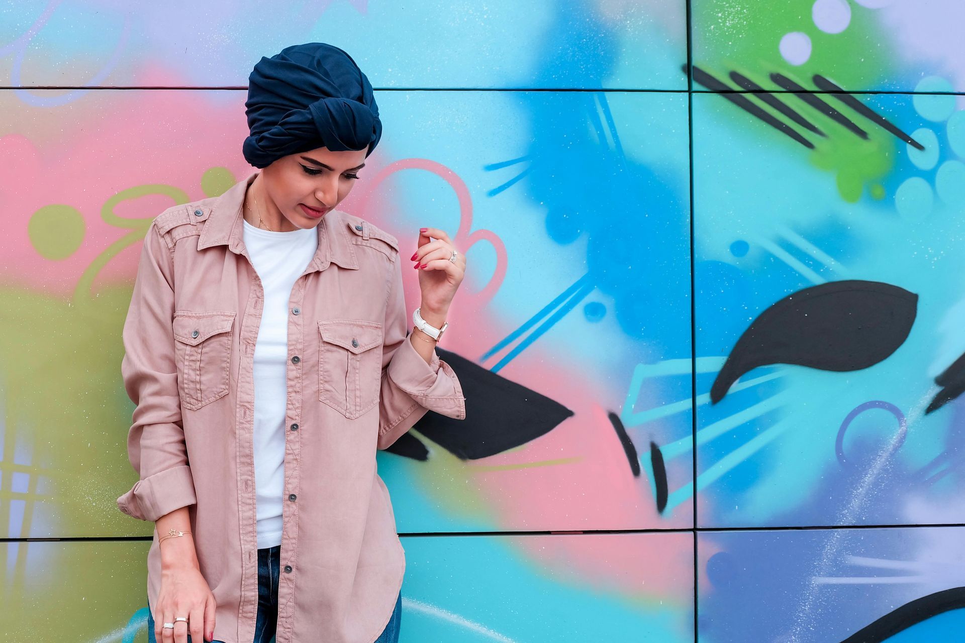 Woman wearing a blue head covering and pink shirt, looking down, leaning against a colorful graffiti wall.