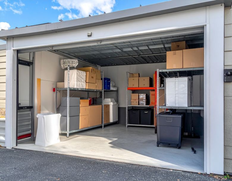 Open storage garage with shelves, boxes, a cabinet, and a trash bin inside.