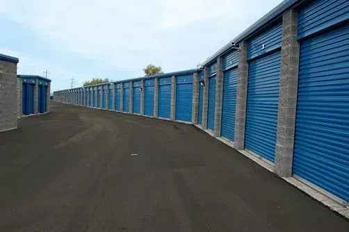 Row of blue storage unit doors along a paved driveway under a cloudy sky