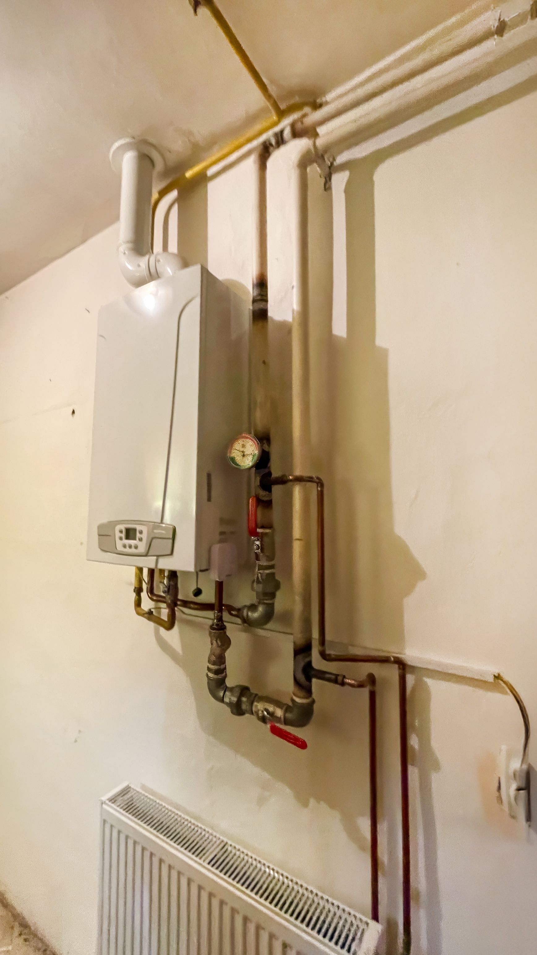 Wall-mounted white boiler and pipes above a radiator in a small utility room.