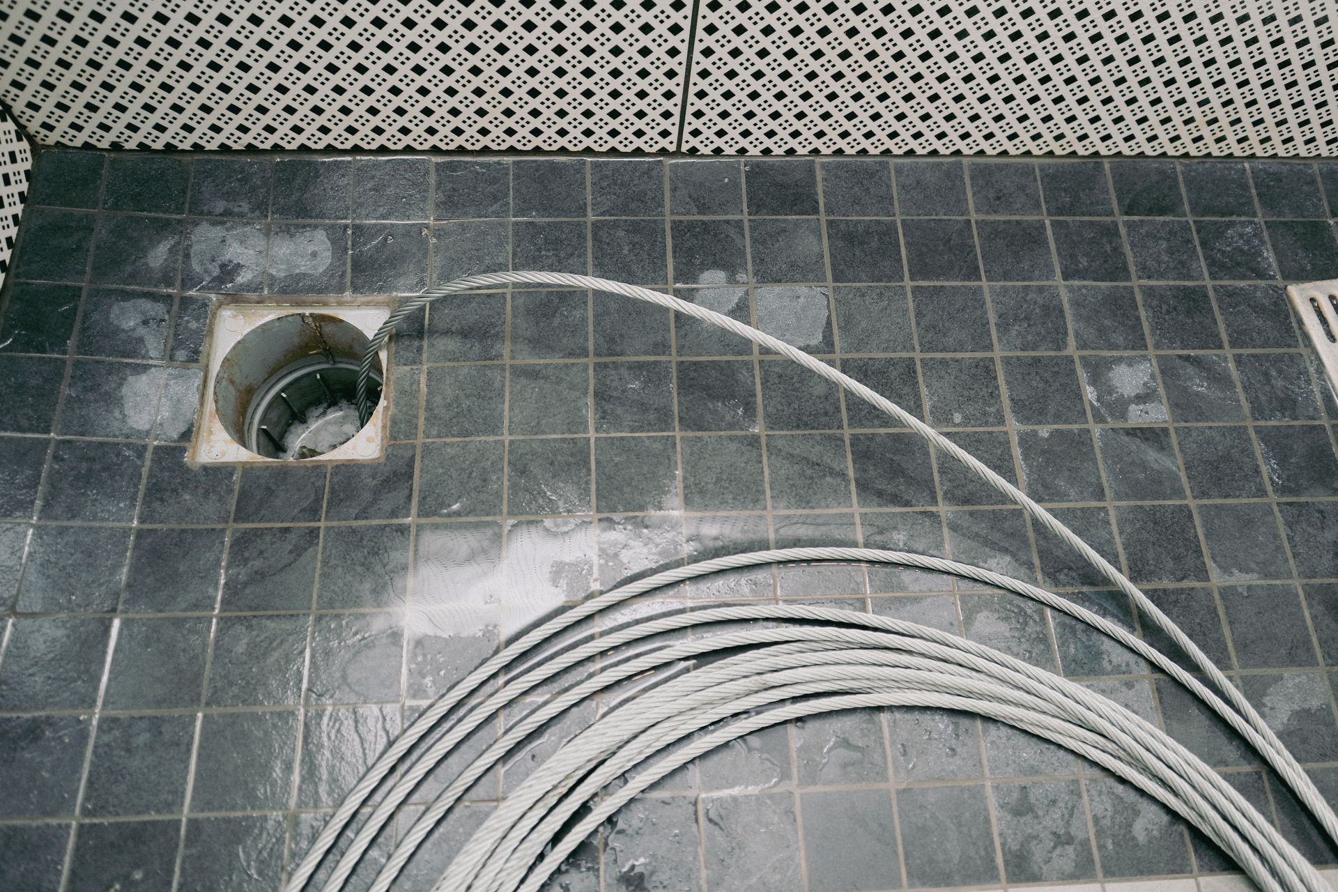 Coiled white cable on a dark tiled floor beside a square drain opening