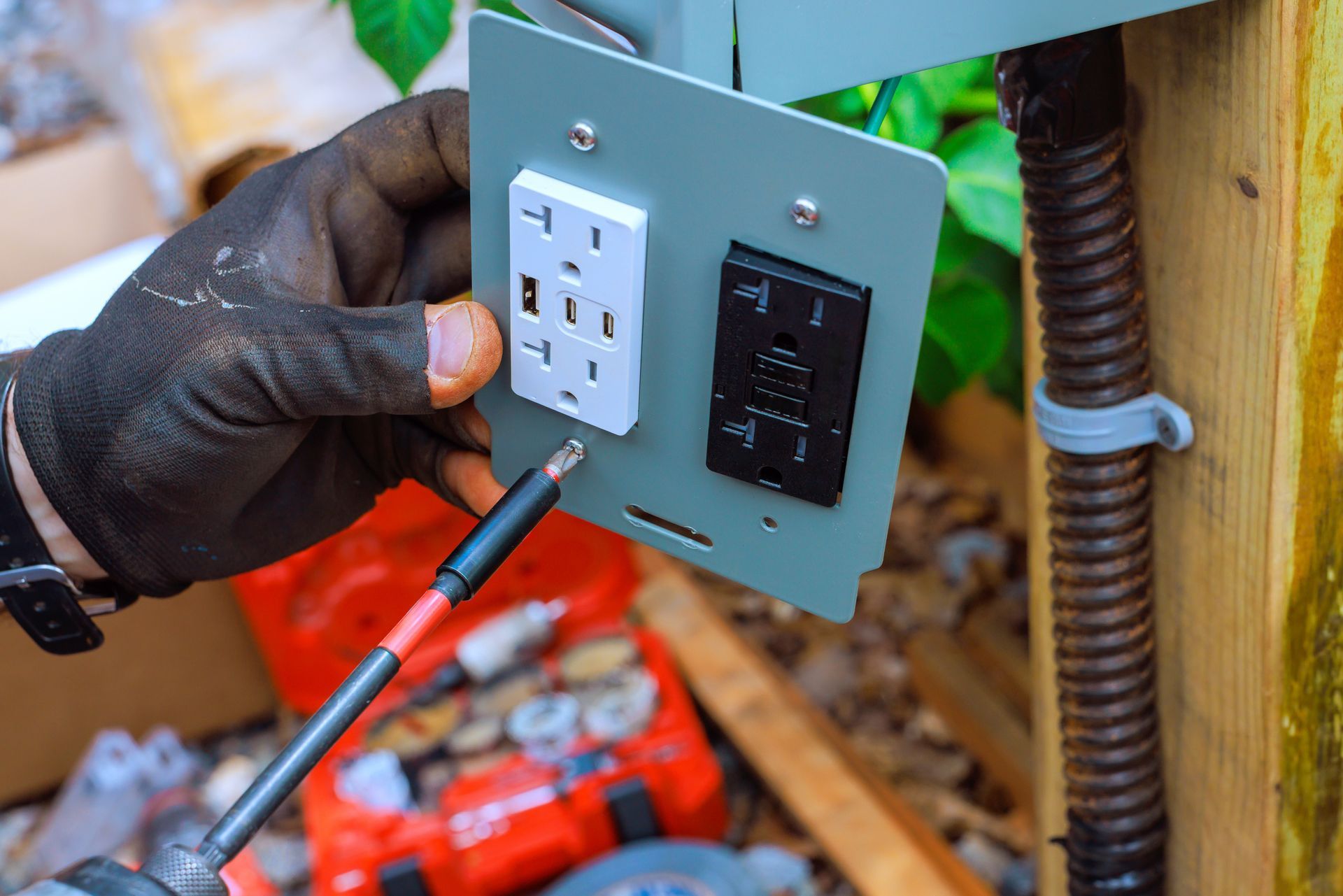 Gloved hand testing a wall outlet with a voltage tester on a metal plate outdoors