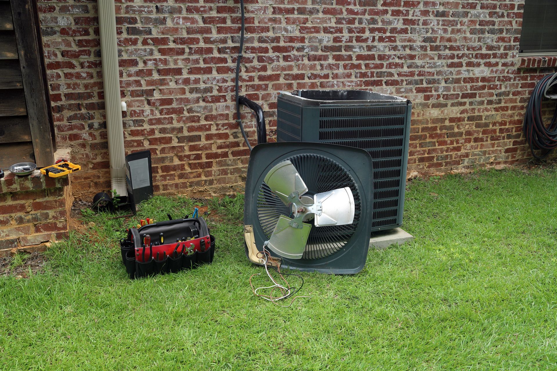 Outdoor HVAC unit beside a brick wall with a detached fan on the grass and a red toolbox nearby