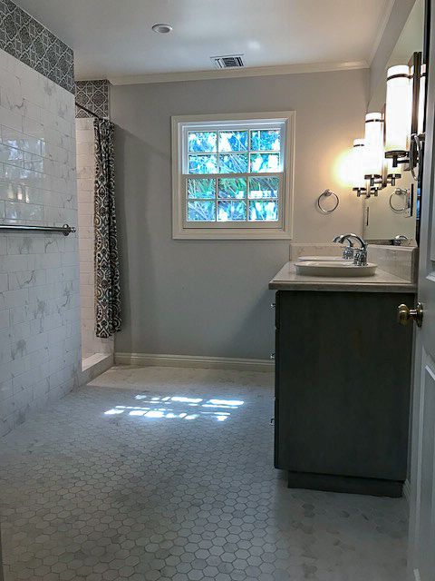 Bright bathroom with tiled shower, vanity sink, wall sconce, and sunlight through a small window.