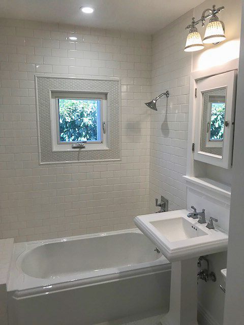White tiled bathroom with a bathtub, sink, mirror, and two windows.
