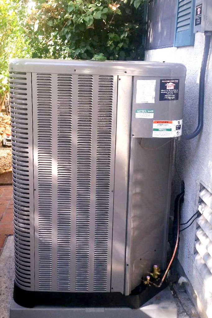 Outdoor air conditioner unit beside a house wall, with a gray grille and metal cabinet.