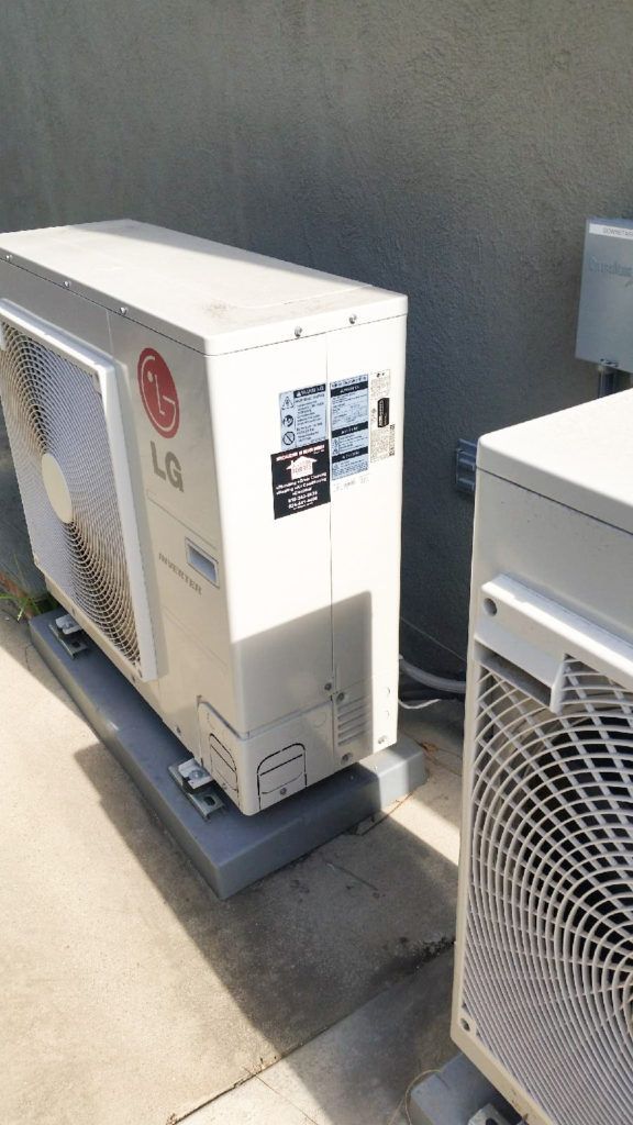 Outdoor HVAC units beside a building wall, including a white unit on a concrete pad and a gray grille unit.