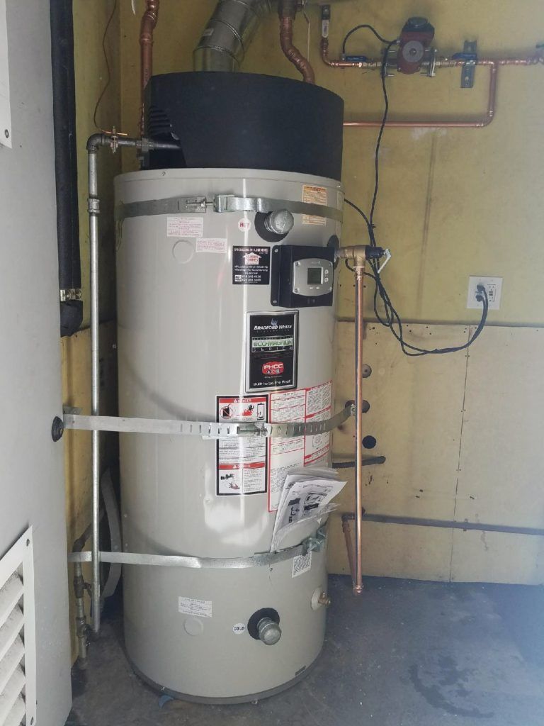 Large beige water heater with warning labels, pipes, and wiring in a utility room against a yellow wall.