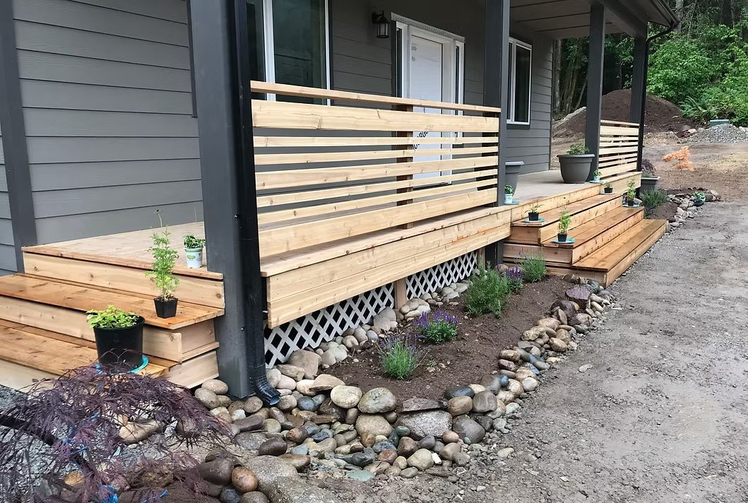 Wooden porch and steps with horizontal railings, surrounded by landscaping and a gravel driveway.