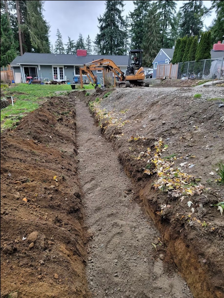 Trench dug in a yard, with a small excavator in the background, near a house.