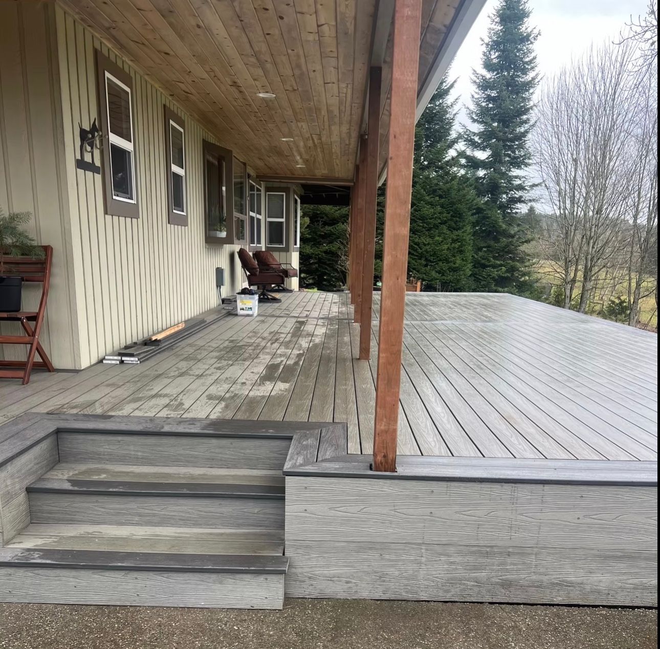 Covered wooden deck with steps leading up to a house. Exterior shot with siding and a glimpse of trees.