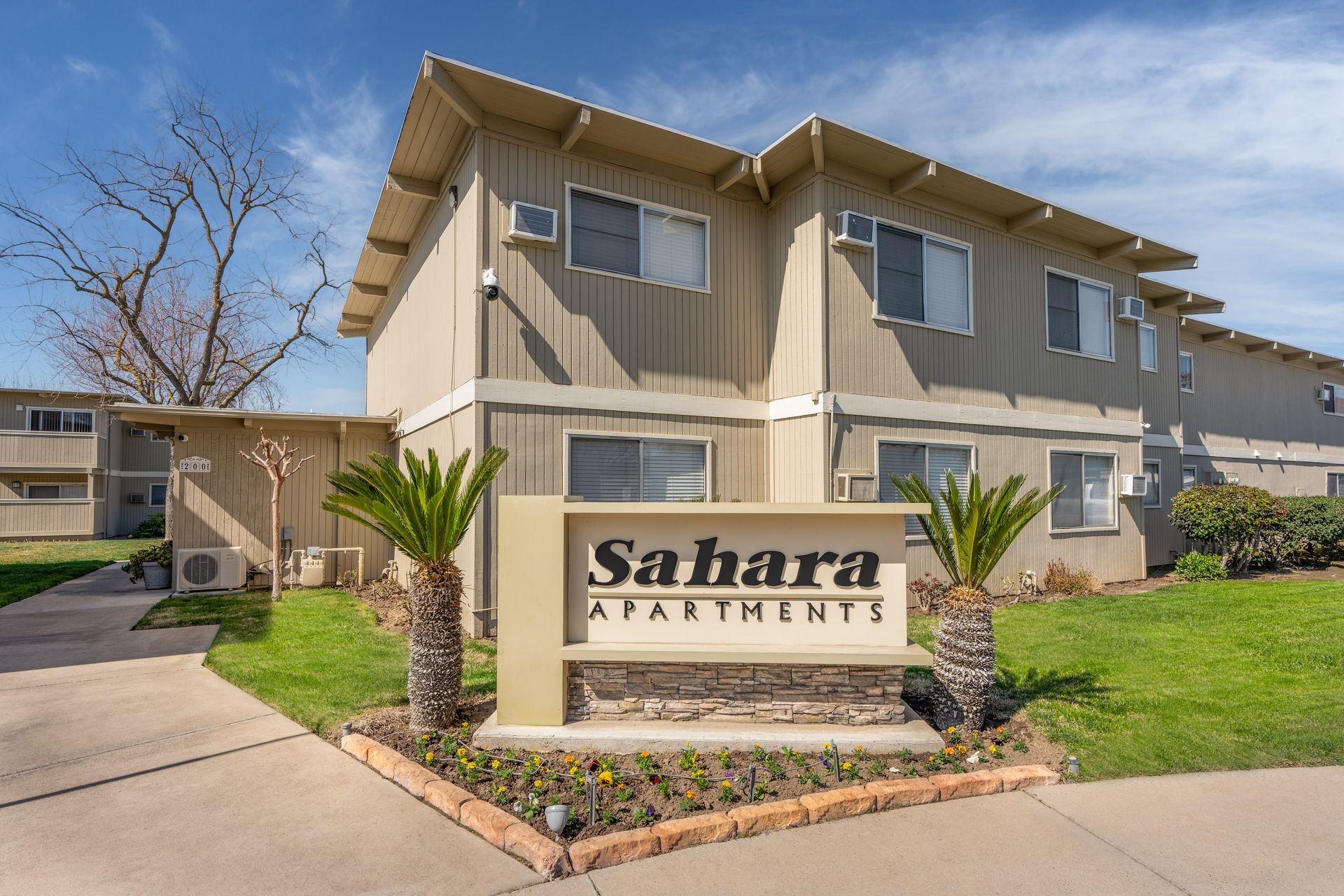 Sahara Apartments sign in front of a two-story beige apartment building. Palm trees flank the sign on a sunny day.