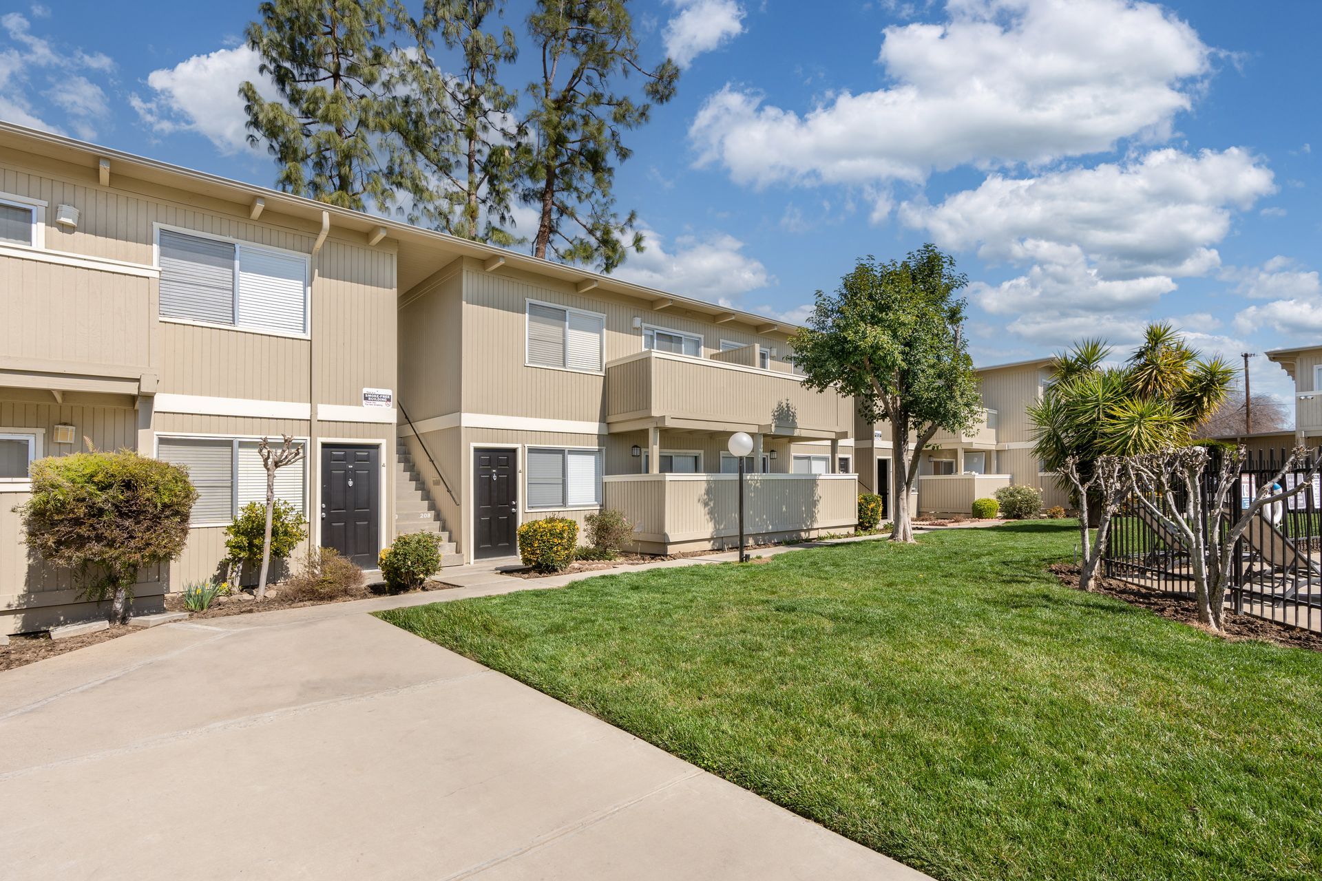 Two-story tan apartments with green lawn, trees, and a blue sky.