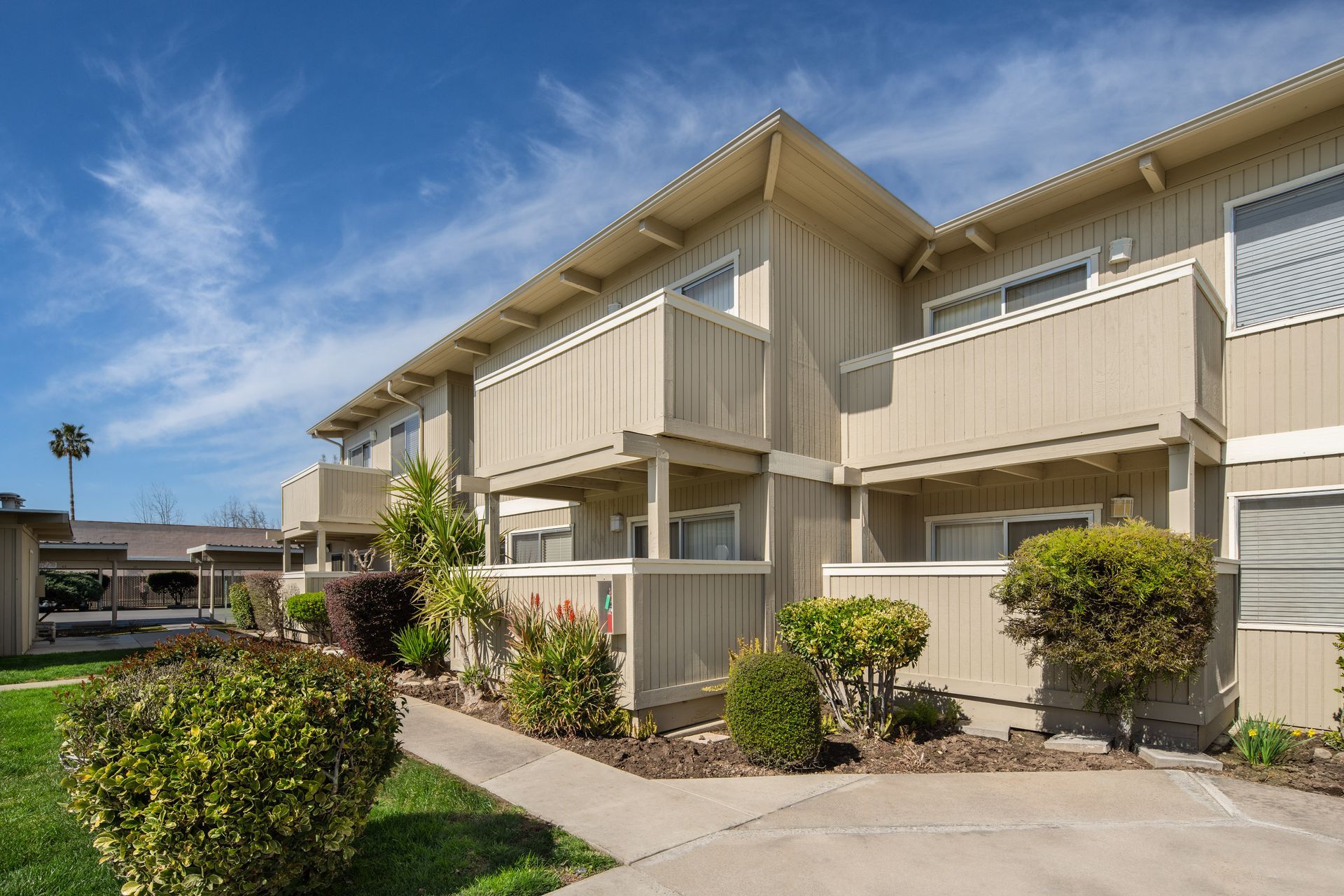 Apartment building with beige siding, small balconies, and manicured landscaping under a blue sky.