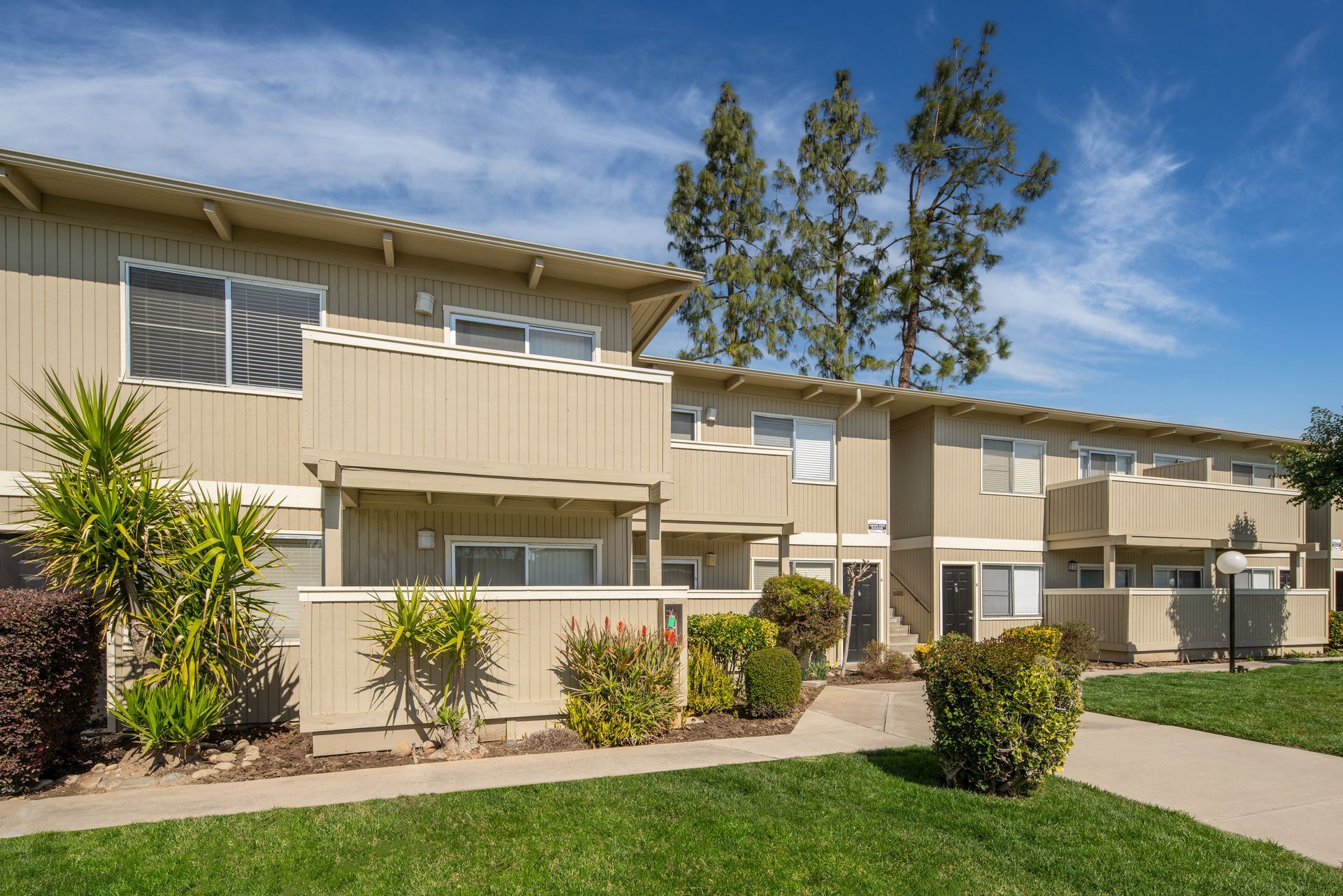 Two-story beige townhomes with white window frames, green lawns, and blue sky.