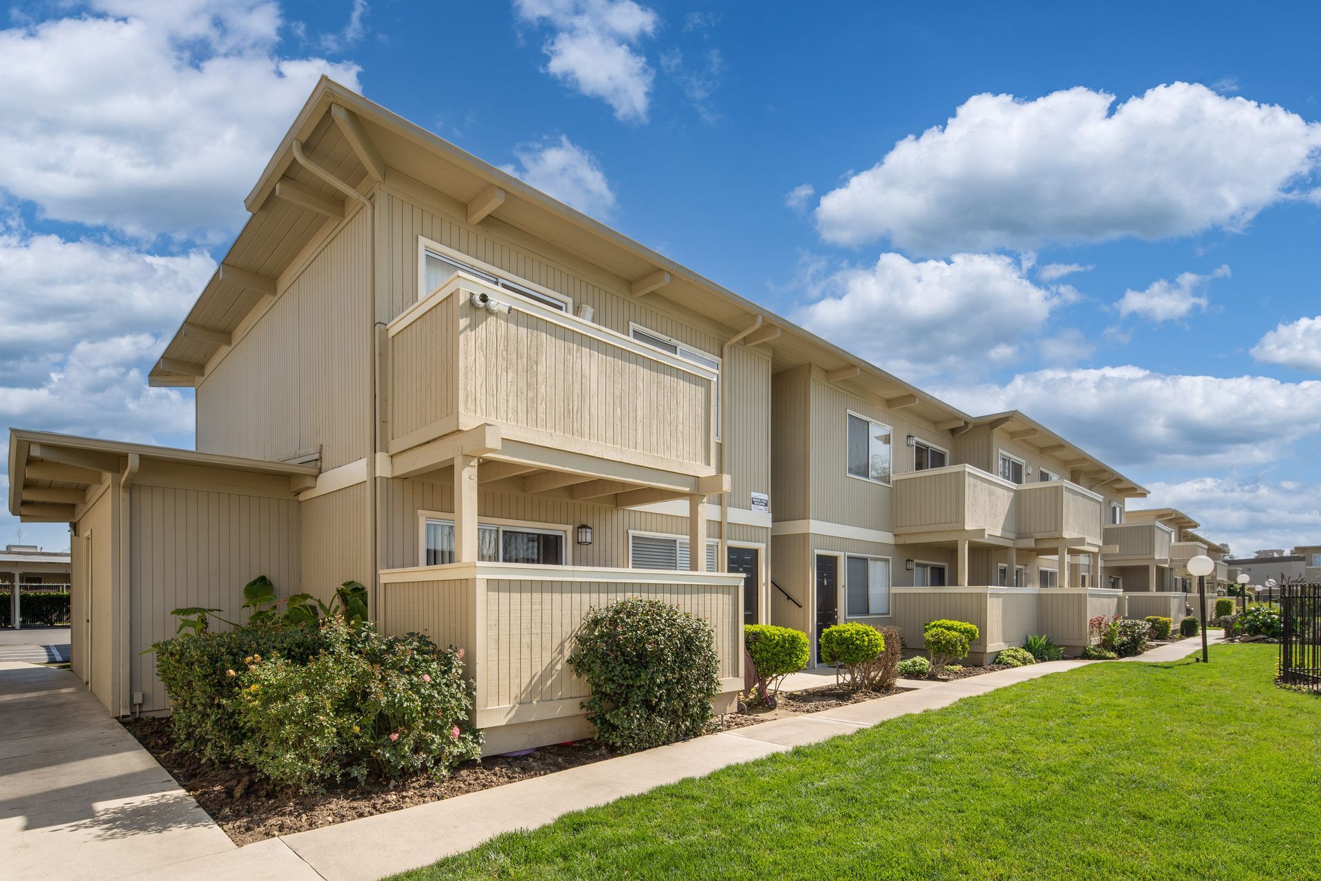 Two-story tan townhouses with balconies and a grassy lawn under a blue sky with clouds.