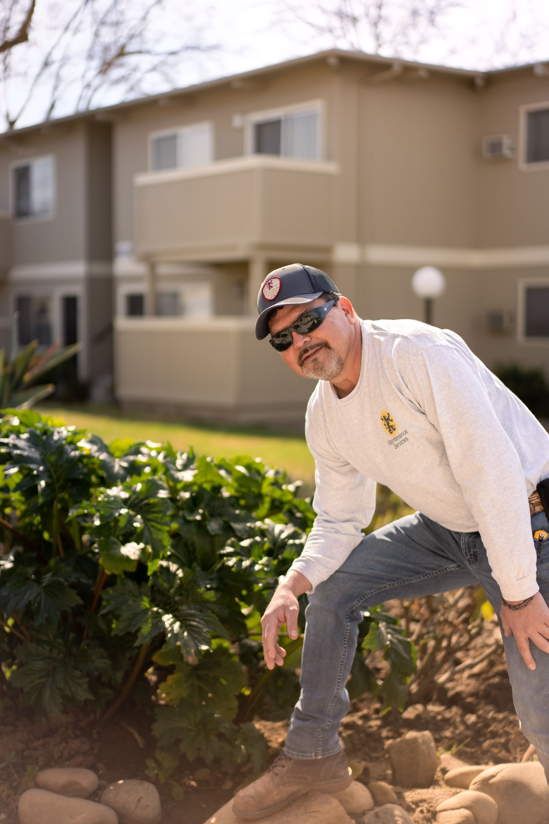 Man in work clothes, sunglasses, and hat leans toward camera near rocks, apartment building background.