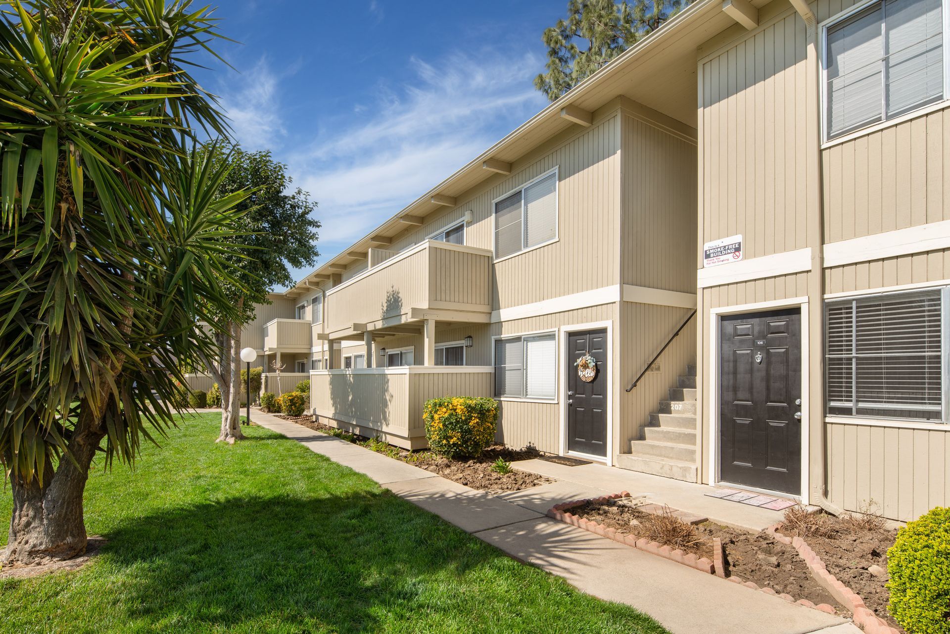 Apartment building with light brown siding, black doors, and green lawn under a blue sky.