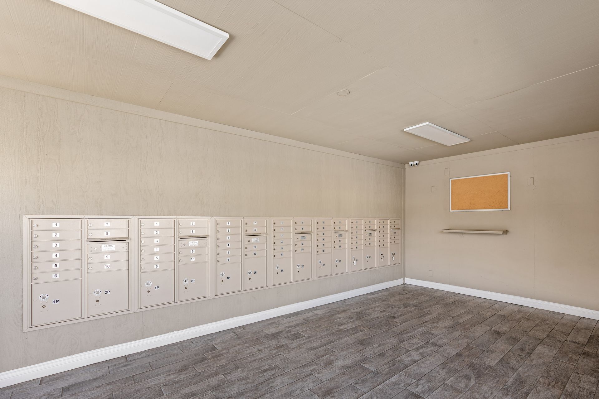 Apartment mailboxes along a wall, next to a bulletin board, in a neutral-toned room.