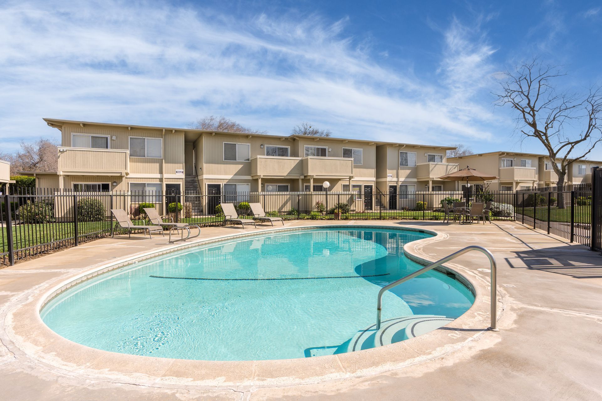 Swimming pool in front of two-story beige apartment buildings with lounge chairs on concrete deck. Blue sky.