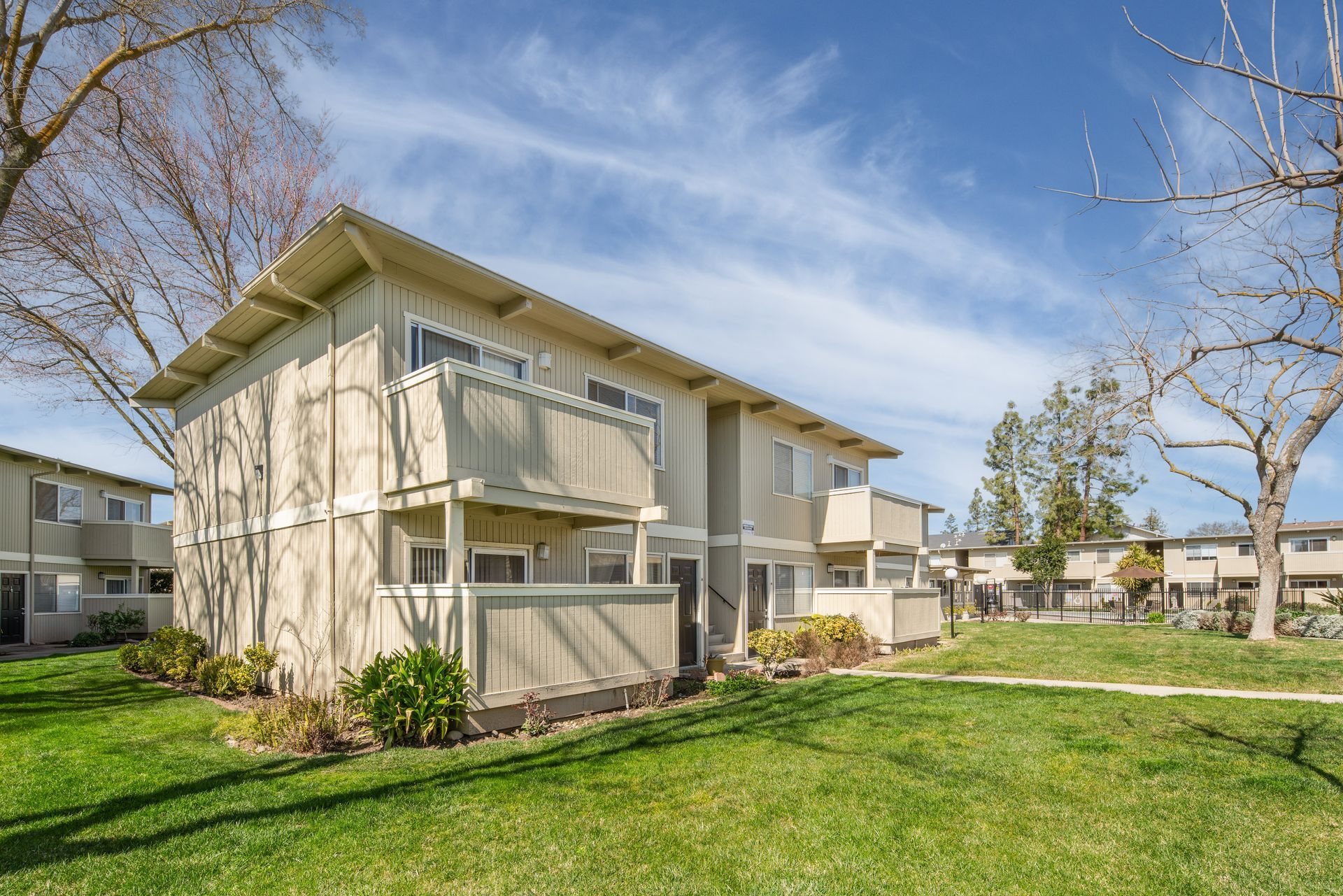 Two-story beige apartment buildings with balconies, green grass lawn, and blue sky.