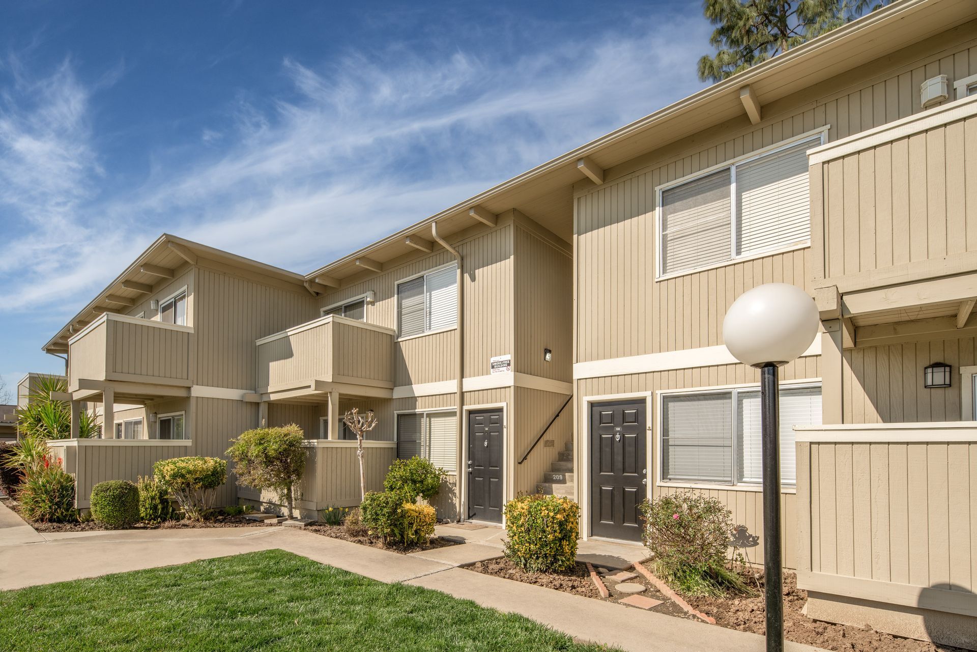 Two-story beige apartment building with balconies and black doors, small bushes in front, blue sky.