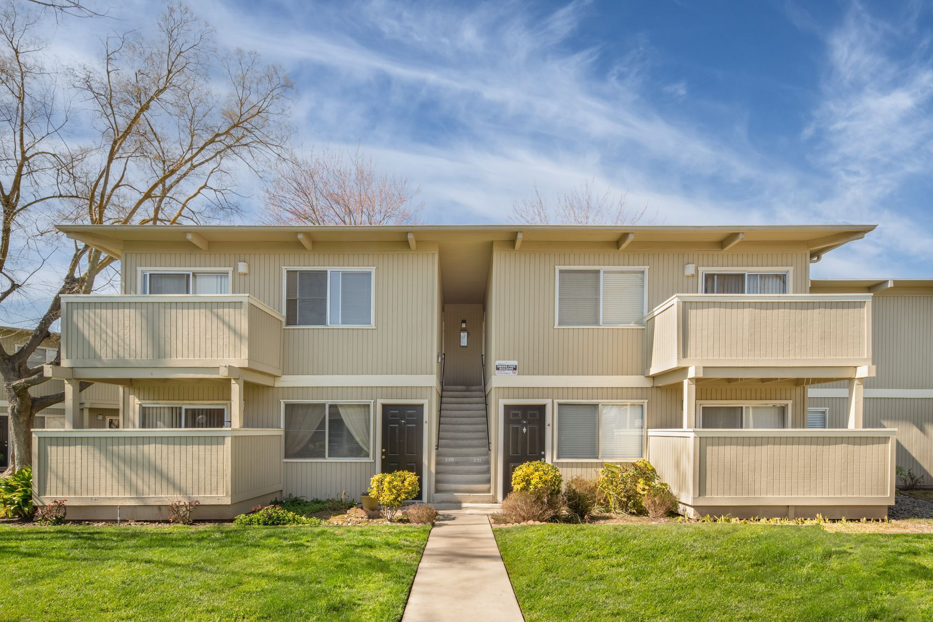 Two-story beige apartment building with balconies, central stairs, and green lawn under a blue sky.