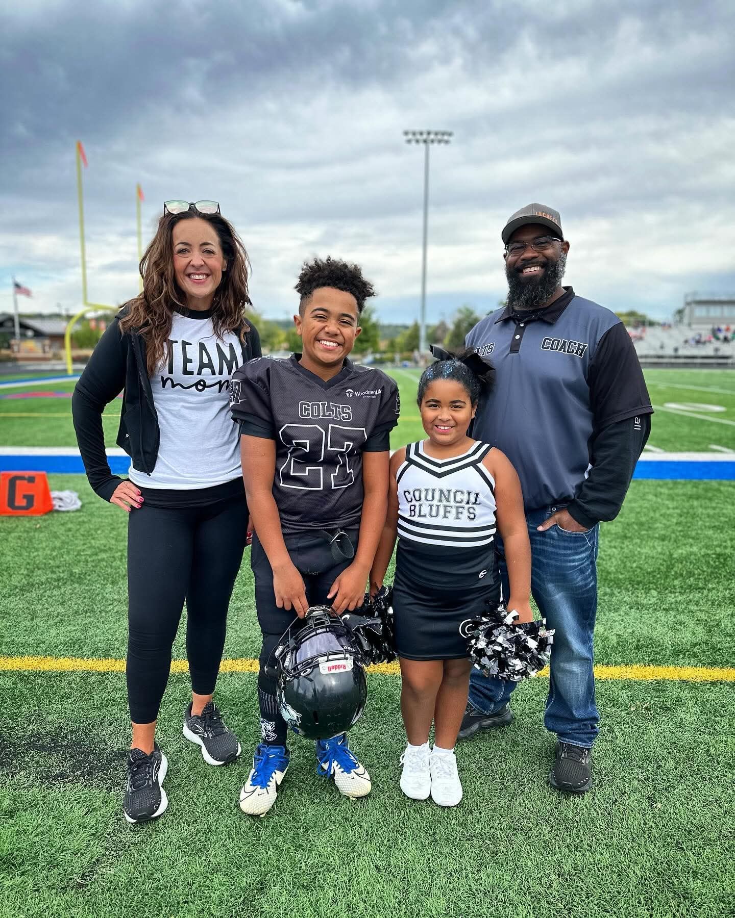 Family of four posing on a football field, smiling with helmets, under cloudy skies.
