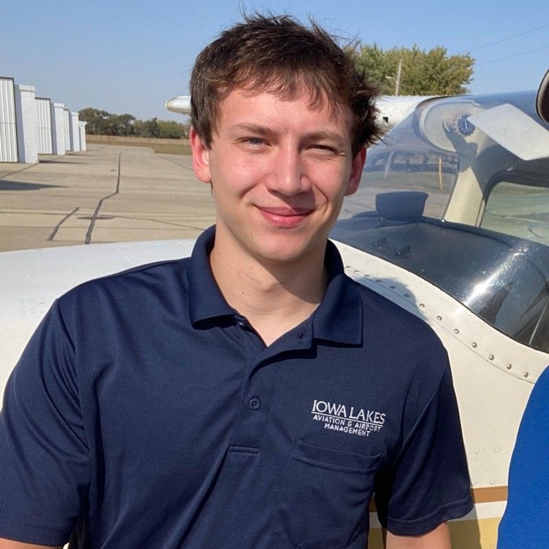 Smiling person in a navy polo standing beside a small airplane on a sunny runway