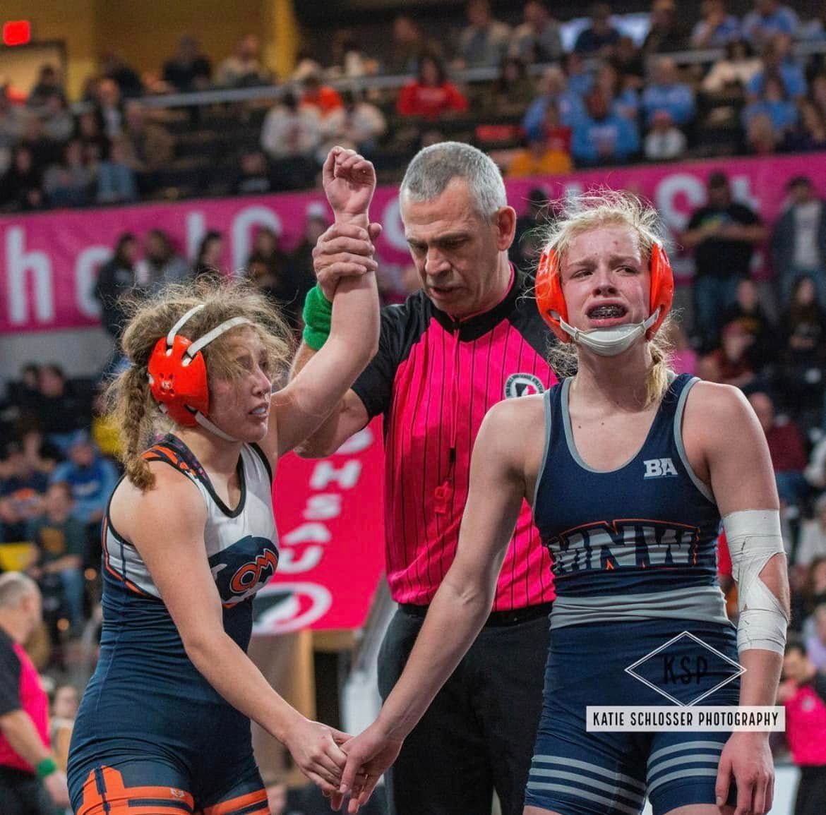 Wrestling referee raises the hand of a blue-singlet wrestler while a red-singlet opponent stands nearby.