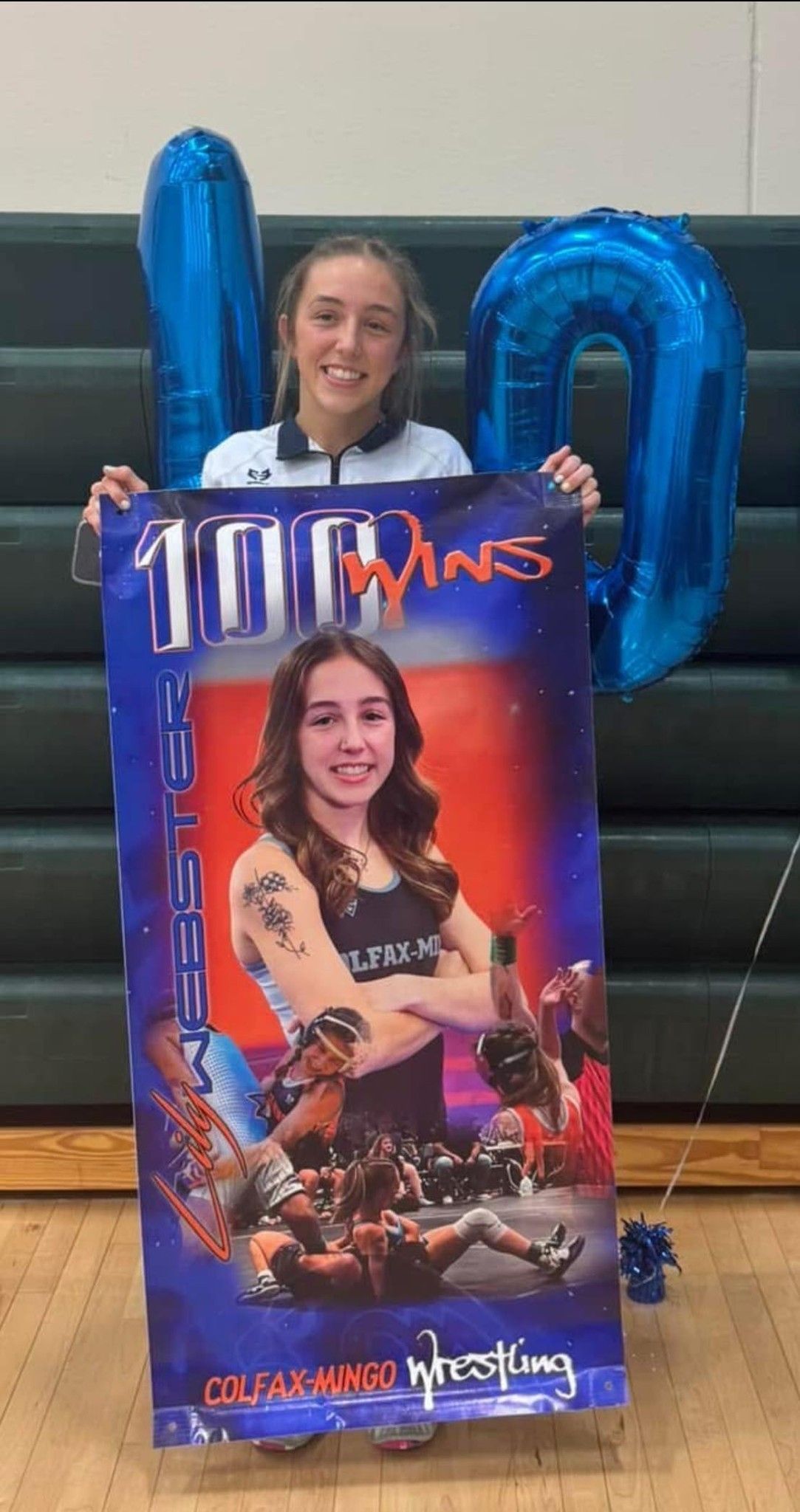 Person in a gym holds a large JoJo Siwa poster with blue balloons behind them.