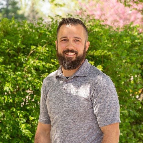 Smiling bearded man in a gray shirt standing outdoors with green and pink flowering bushes behind him